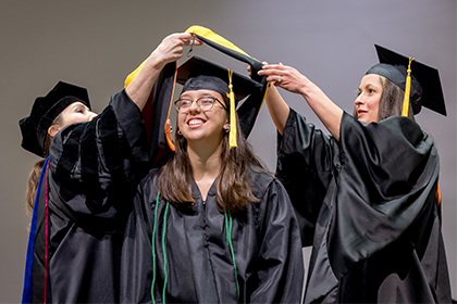 3 women in graduation caps and gowns, 2 placing sash over the head of the women in the center